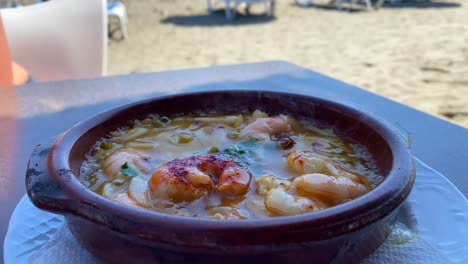 Traditional-hot-sizzling-Spanish-Gambas-pil-pil-in-a-restaurant-at-the-beach-in-Marbella-Spain,-delicious-spicy-shrimp-dish-with-garlic,-sea-view,-4K-shot
