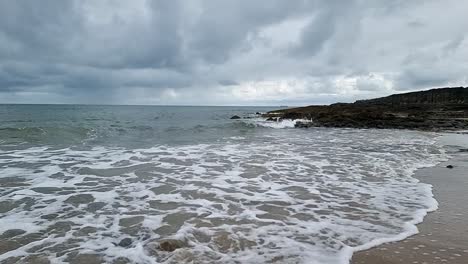 Schäumende-Meereswellen-In-Zeitlupe,-Die-über-Die-Sandige,-Zerklüftete-Walisische-Strandküste-Spülen