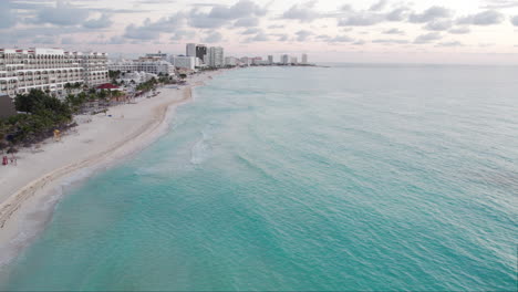 Aerial-Drone-shot-of-rolling-waves-in-a-crystal-clear-blue-ocean-in-a-white-sand-beach-with-hotels-and-resorts-in-Cancun,-Mexico