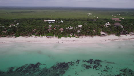 Aerial-Drone-shot-of-cabins-and-huts-surrounded-by-palm-trees-in-front-of-a-white-sand-beach-and-a-crystal-clear-blue-ocean-in-Tulum,-Mexico