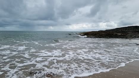 Slow-motion-foaming-ocean-waves-washing-over-sandy-rugged-Welsh-beach-coastline