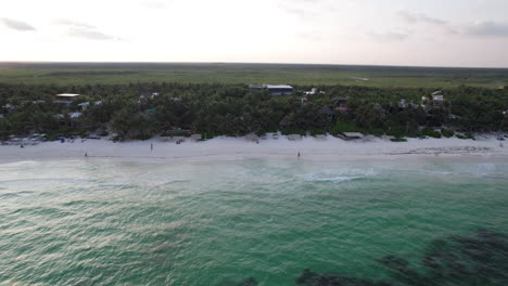 Aerial-Drone-shot-of-cabins-and-huts-surrounded-by-palm-trees-in-a-white-sand-beach-with-crystal-clear-blue-ocean-in-Tulum,-Mexico