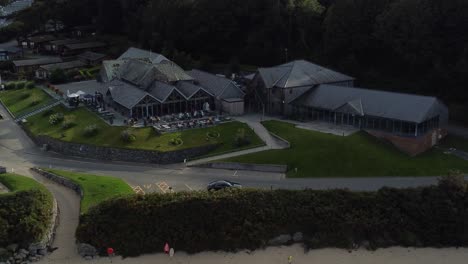 Establishing-aerial-view-over-old-fashioned-tavern-style-beachfront-exclusive-restaurant-shaded-by-Welsh-mountainside-on-secluded-beach