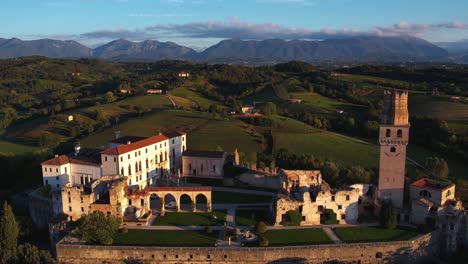 Aerial-view-of-a-medieval-castle-on-top-of-a-hill,-surrounded-by-prosecco-vineyards,-in-Italy,-with-mountains-in-the-background