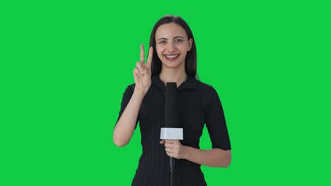 Happy-Indian-female-news-reporter-showing-victory-sign-Green-screen