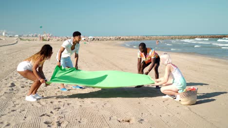 Diverse-men-and-women-laying-blanket-on-beach
