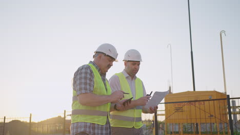 Two-engineers-discussing-project-on-a-construction-site-a-worker-wearing-a-helmet-during-the-sunset