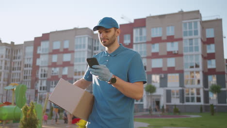 The-postman-with-glasses-carries-the-parcel-and-looks-at-the-delivery-address-via-mobile-phone.-search-for-the-address-of-the-delivery-customer.-Delivery-guy-with-a-cap-and-a-box