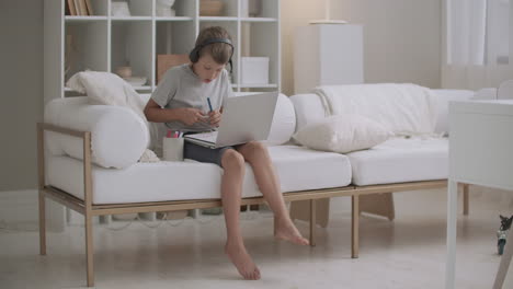 Ein-Kleiner-Junge-Hört-Musik-über-Kopfhörer-Mit-Einem-Laptop-Und-Zeichnet-Bilder,-Die-Zu-Hause-Am-Tisch-Sitzen
