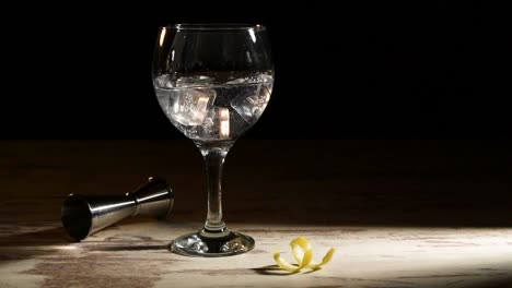 Person-pouring-ice-cubes-into-glass-of-gin-tonic-with-lemon-and-jigger-on-table-in-dark-room