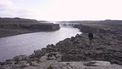 Traveler-walking-along-rocky-shore-of-river-towards-waterfall