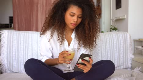 Ethnic-woman-drinking-juice-and-browsing-smartphone-at-home