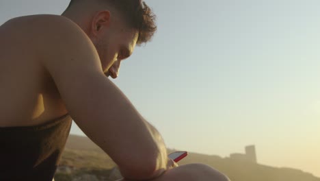 Man-sitting-on-seashore-and-browsing-smartphone