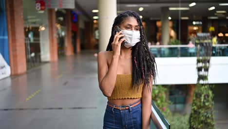 Young-black-woman-in-face-mask-speaks-on-smart-phone-in-commercial-mall