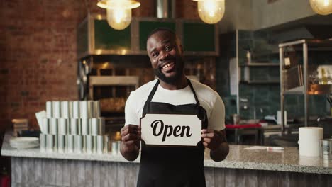 A-Black-person-in-a-black-apron-holds-a-sign-Open-in-his-hands.-The-guy-smiles-and-looks-at-the-camera.-Video-filmed-in-high-quality