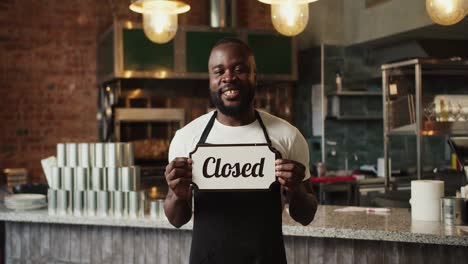 Un-Tipo-Negro-Con-Un-Delantal-Negro-Sostiene-Un-Cartel-Cercano-En-Sus-Manos.-El-Establecimiento-Está-Cerrado.-Vídeo-Filmado-En-Alta-Calidad