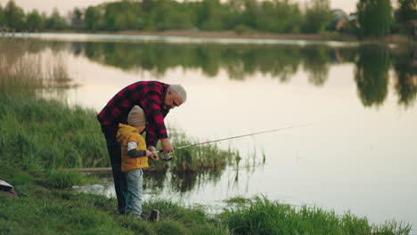 happy-grandfather-and-little-grandson-are-fishing-together-in-summer-morning-standing-on-beautiful-shore-of-lake