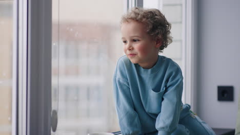 chubby-slavic-little-boy-with-charming-curly-hair-is-sitting-on-windowsill-in-room-in-snowy-day-looking-out-window