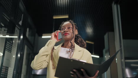 Mujer-De-Negocios-Africana-Hablando-De-Teléfono-Inteligente-En-El-Interior-De-La-Oficina.-Empresaria-Negra-Sonriente-Llamando-A-Un-Teléfono-Móvil-En-El-Pasillo.-Retrato-De-Un-Amigable-Afroamericano-Caminando-En-El-Pasillo-Del-Centro-De-Negocios.