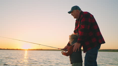 Alter-Fischer-Bringt-Seinem-Enkel-Bei,-Fische-Zu-Fangen,-Indem-Er-Angelt---Glückliche-Familie-In-Der-Natur,-Die-Am-Fluss-Angelt