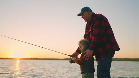 old-fisher-and-little-grandson-are-catching-fish-by-rod-in-river-or-lake-shore-rest-in-nature-in-evening