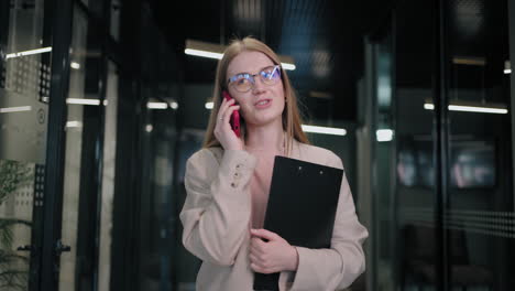 Portrait-of-a-smiling-Business-woman-with-glasses-walking-in-the-hallway-of-the-office-of-a-private-and-talking-on-the-phone-with-customers