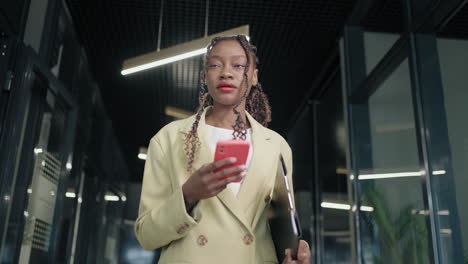 Retrato-De-Una-Mujer-De-Negocios-Negra-Caminando-Por-El-Pasillo-De-Un-Centro-De-Negocios-Y-Escribiendo-Un-Mensaje-En-Su-Teléfono