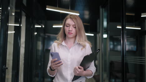 A-blonde-business-woman-walks-down-the-corridor-and-writes-a-message-and-looks-at-the-phone-screen