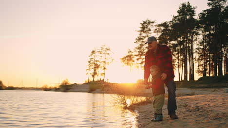 granddad-and-grandson-are-resting-on-shore-of-river-and-fishing-in-sunset-time-hiking-in-nature-in-summer