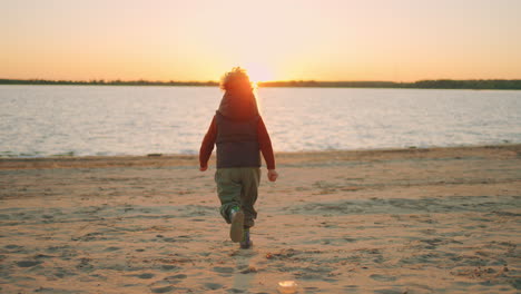 Un-Niño-Alegre-Y-Rizado-Corre-Sobre-La-Costa-Arenosa-Del-Río-Al-Atardecer,-Vista-Trasera-En-Cámara-Lenta-Después-Del-Disparo