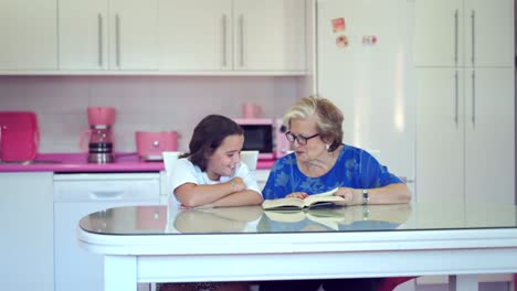 Elderly-woman-reading-book-with-granddaughter-in-light-kitchen-at-home