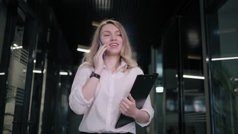 Smiling-businesswoman-looking-on-cellphone-indoors.-Surprised-business-woman-reading-message-on-mobile-phone-in-office-corridor.-woman-looking-smartphone-screen-in-business-center.-walking-office