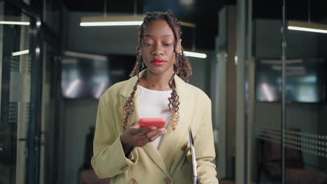 Retrato-De-Una-Mujer-De-Negocios-Negra-Caminando-Y-Hojeando-Las-Noticias-Por-Teléfono-En-El-Pasillo-De-La-Oficina