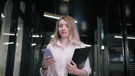 Smiling-businesswoman-looking-on-cellphone-indoors.-Surprised-business-woman-reading-message-on-mobile-phone-in-office-corridor.-woman-looking-smartphone-screen-in-business-center.-walking-office