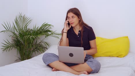 Woman-speaking-on-mobile-phone-and-using-laptop-in-bedroom