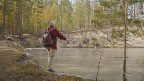grey-haired-man-dressed-red-checkered-shirt-is-casting-fishing-rod-in-river-relaxing-on-shore-of-reservoir