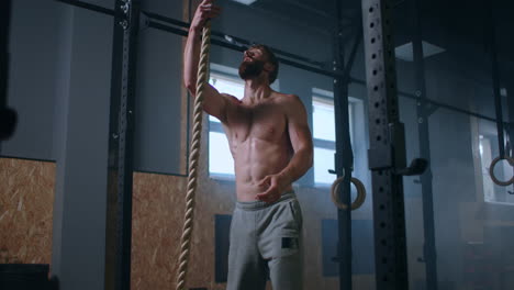 a-man-in-the-gym-climbs-up-the-rope-to-the-top.-perseverance-and-strength.-a-sweaty-man-without-a-t-shirt-is-training.
