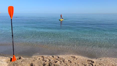 Girl-puddle-surfing-in-the-sea-in-Nerja-Malaga,-stand-up-puddle-boarding-on-a-sunny-day-in-Spain,-fun-vacation-activity,-adventurous-water-sport,-4K-static-shot