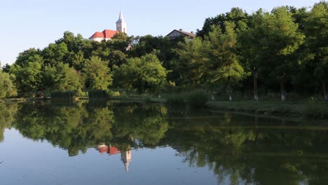 Church-steeple-behind-grove-of-trees-reflecting-off-surface-of-calm,-scenic-pond