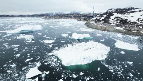 Floating-iceberg-in-deep-ocean-with-small-town-in-background,-aerial-view