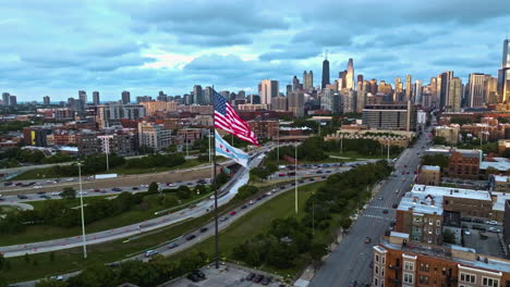 Vista-Aérea-Alrededor-De-Nosotros-Y-La-Bandera-De-Chicago-Con-Fondo-De-Horizonte-Iluminado-Por-El-Sol