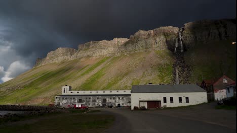 Toma-Panorámica-De-La-Bahía-De-Djúpavík-Con-Montañas,-Cascada-Y-Antigua-Fábrica-Durante-El-Cielo-Gris-Nublado-Al-Atardecer,-Islandia