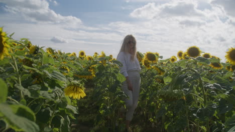 Blonde-woman-in-sunglasses-walks-through-sunflowers,-frontal-slomo