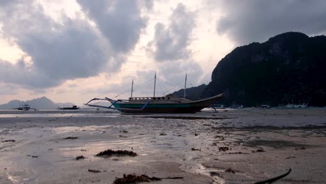 Traditional-outrigger-tour-boat-moored-on-shoreline-during-low-tide-with-dramatic-cloudy-twilight-sunset-in-El-Nido,-Palawan-in-the-Philippines,-Southeast-Asia