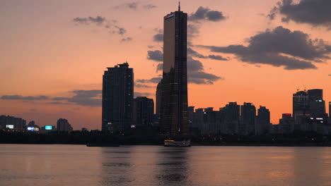 63-Building-In-Yeouido-and-Han-River-Sunset-Cruise-Ship-or-Ferry-Travels-Along-Riverbank-in-Urban-Background---Real-Time-Landscape