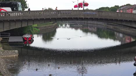 Barco-Turístico-Atracado-Cerca-Del-Puente-En-El-Centro-De-La-Ciudad-De-Kilkenny-Con-Autobús-Y-Gente-Cruzando-El-Puente-En-Verano.