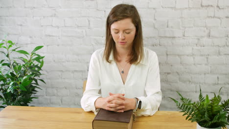 A-young-woman-praying-with-a-bible-and-fingers-clasped-together-in-prayer-sitting-at-a-table