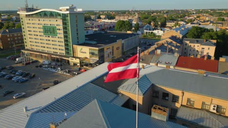 Drone-orbits-around-The-Latvian-national-flag-on-top-of-the-Unity-House-building-at-sunset,-Daugavpils,-Latvia