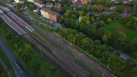 aerial-view-of-a-houses-in-neighbourhood-and-red-train-passing-by-on-tracks-next-to-a-road-where-cars-are-driving-there-is-parking-spots-and-a-small-lake-in-a-location-filled-with-trees-and-gardens
