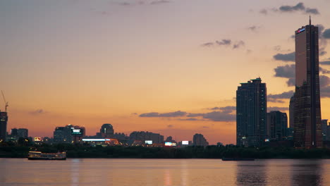 63-Gebäude,-Das-Kreuzfahrtschiff-Han-River-Sunset-Fährt-In-Einem-Atemberaubenden-Orangefarbenen-Himmel,-Der-Sich-Im-Plätschernden-Wasser-Spiegelt